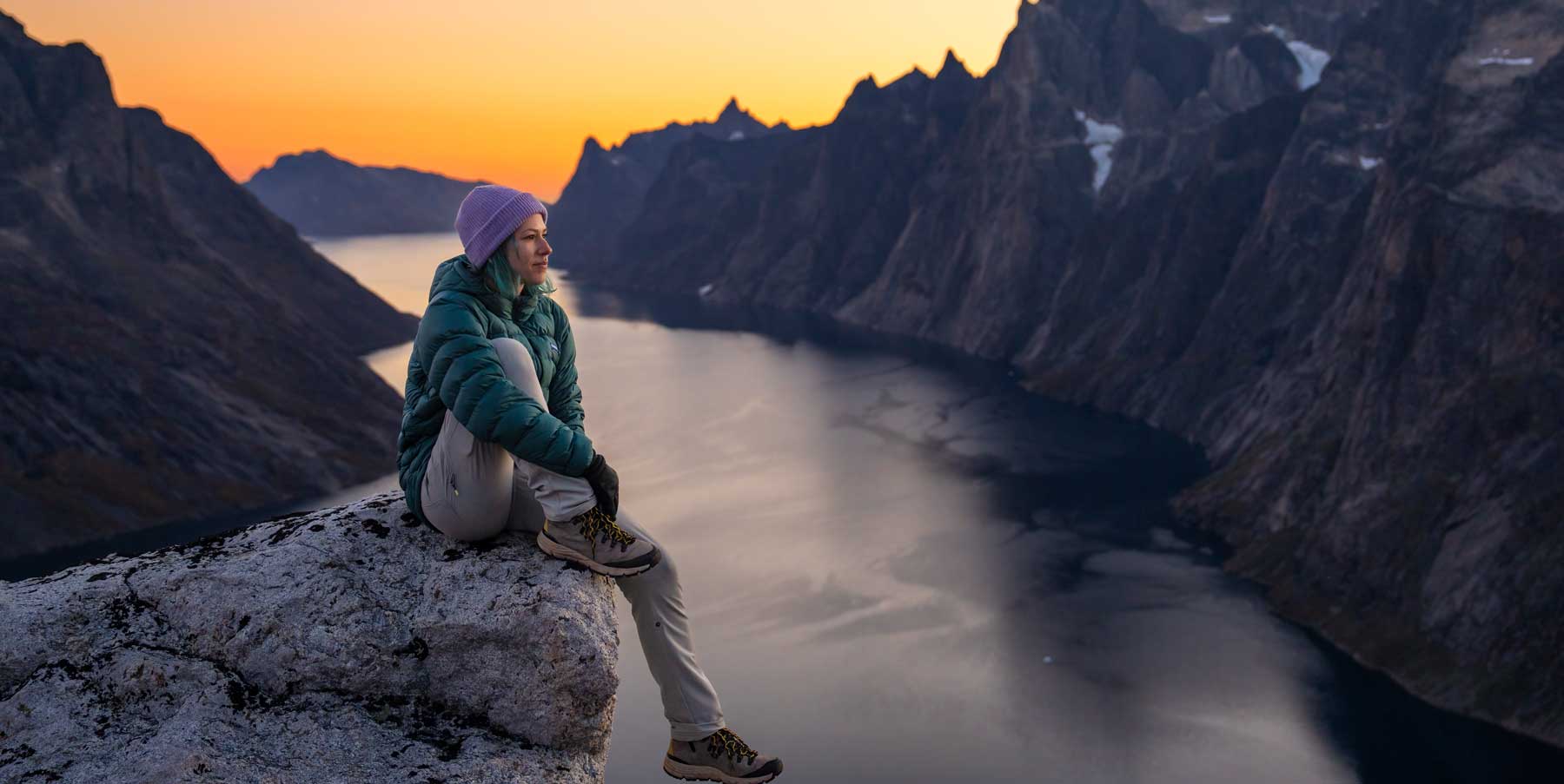 Woman sitting above a rocky fjord at sunset with a teal Eos Jacket